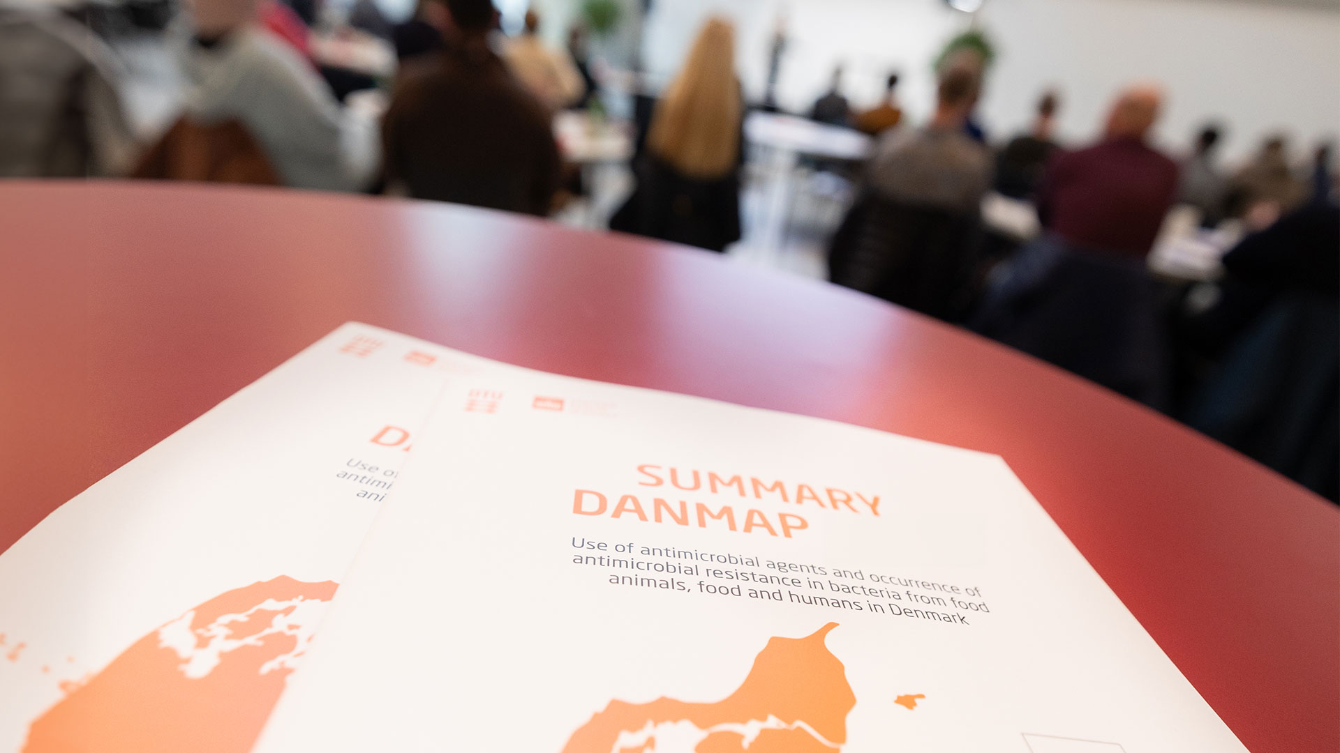 Foto af konferencebord med DANMAP-rapport plus mennesker i baggrunden